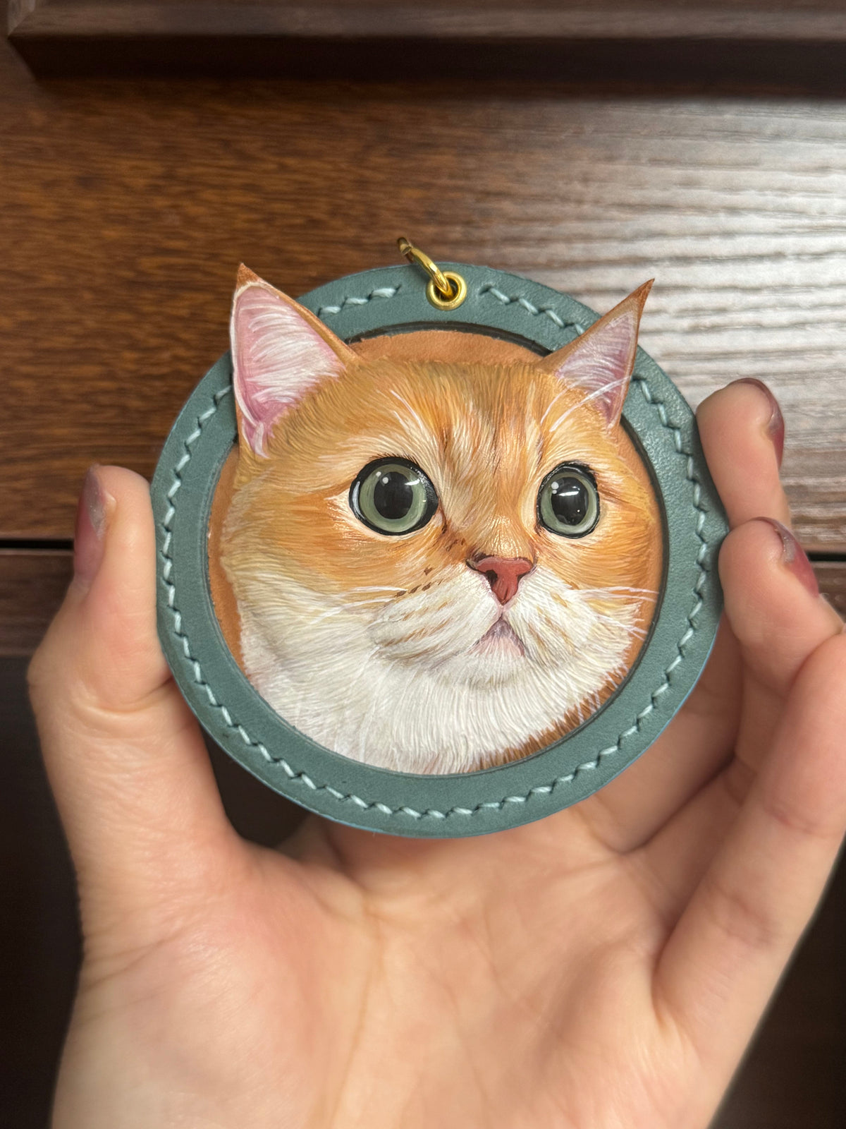 Teal round cat-face figurine keychain with stitched edges and small gold top ring